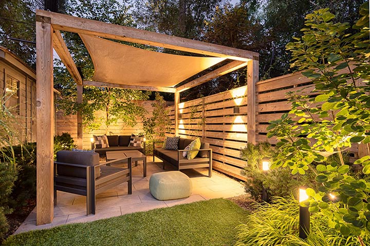 Inviting backyard with a wooden tall privacy fence all around, beautiful modern lighting, and modern black furniture under a wooden pergola.