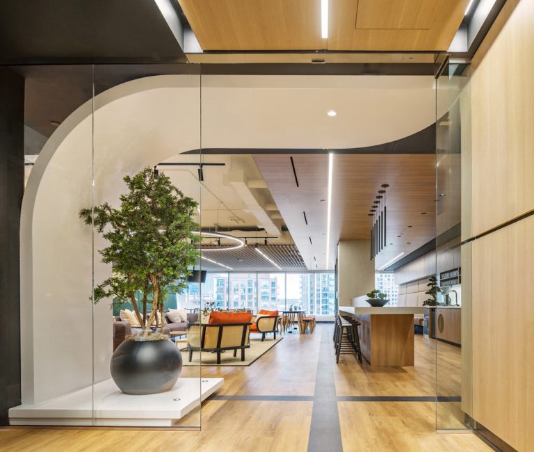 Entrance to a common area of the space which includes a bar on the right with kitchen and on the right there is a lounge area. Open sightlines all the way to the other end of the space looking out the large glass wall and view of buildings