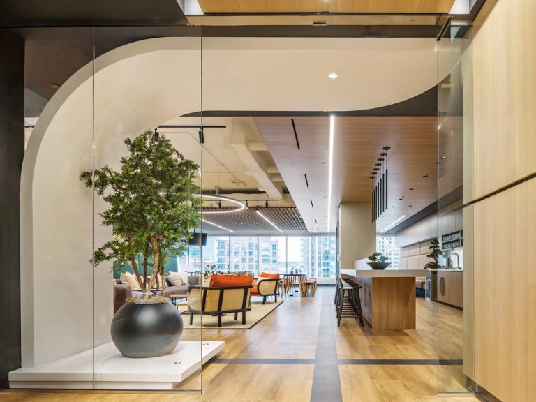 Entrance to a common area of the space which includes a bar on the right with kitchen and on the right there is a lounge area. Open sightlines all the way to the other end of the space looking out the large glass wall and view of buildings