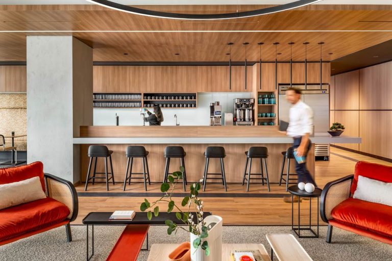 Wood finished kitchen and bar with black bar stools and thin liner lights in black finish above the bar. In the foreground, bamboo lounge chairs with bright red upholstery on the seating.