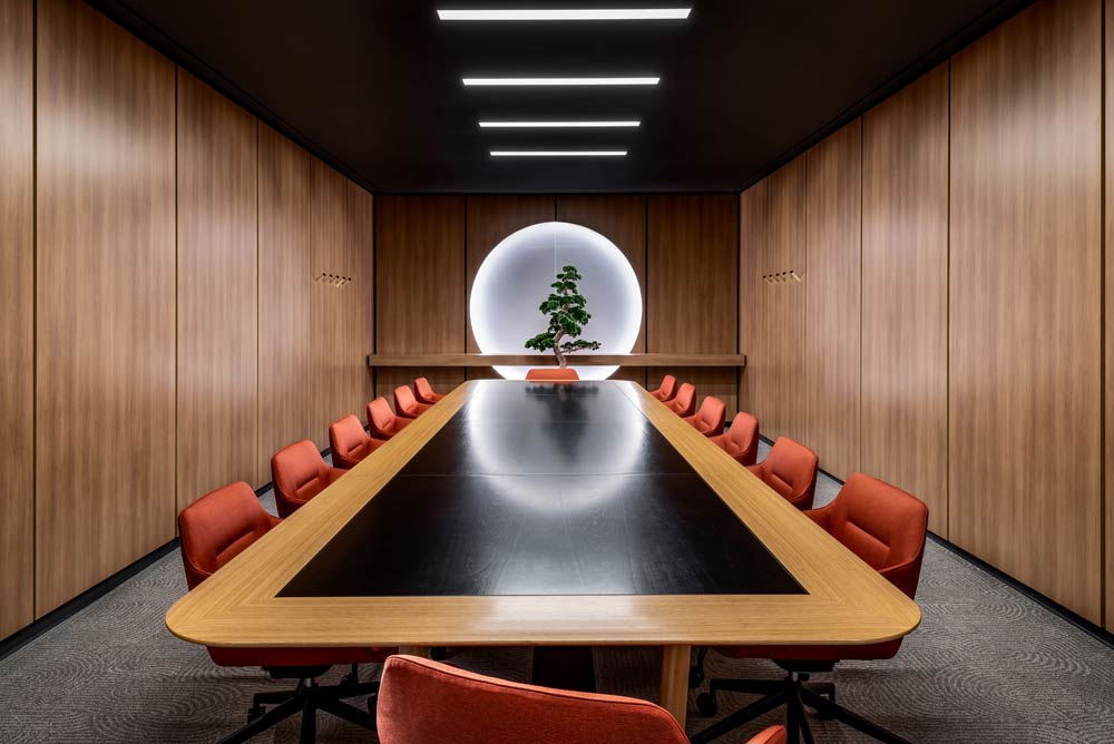 A board room with walls covered in wood finish paneling, a long table with red chairs and a bonsai tree at the end of the table highlighted with a circular light in the wall behind the tree.