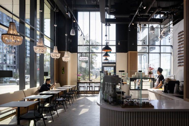 A view from the interior looking at the coffee shop counter toward the street. Across the counter is a seating area with black chairs and elegant chandeliers above.