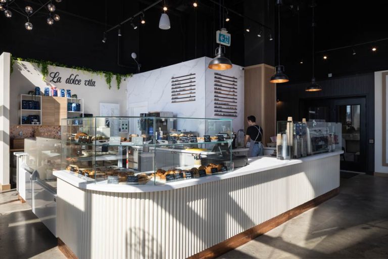 A view at the entire coffee shop area with a fluted counter with rounded corner that stores sweets, pastries and coffee supplies on top, and industrial black pendant lights. The marble stone back wall behind the counter displays the menu in an industrial style.