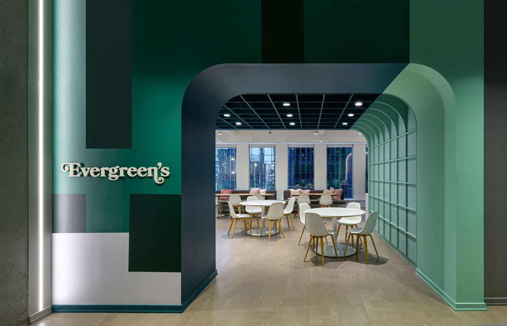 The entrance to 'Evergreens' lounge/bar, the colours on the entrance wall are in different shades of green that create a geometric patters, leading the view inside where white tables and chairs await, and large floor to ceiling windows at the opposite end of the space