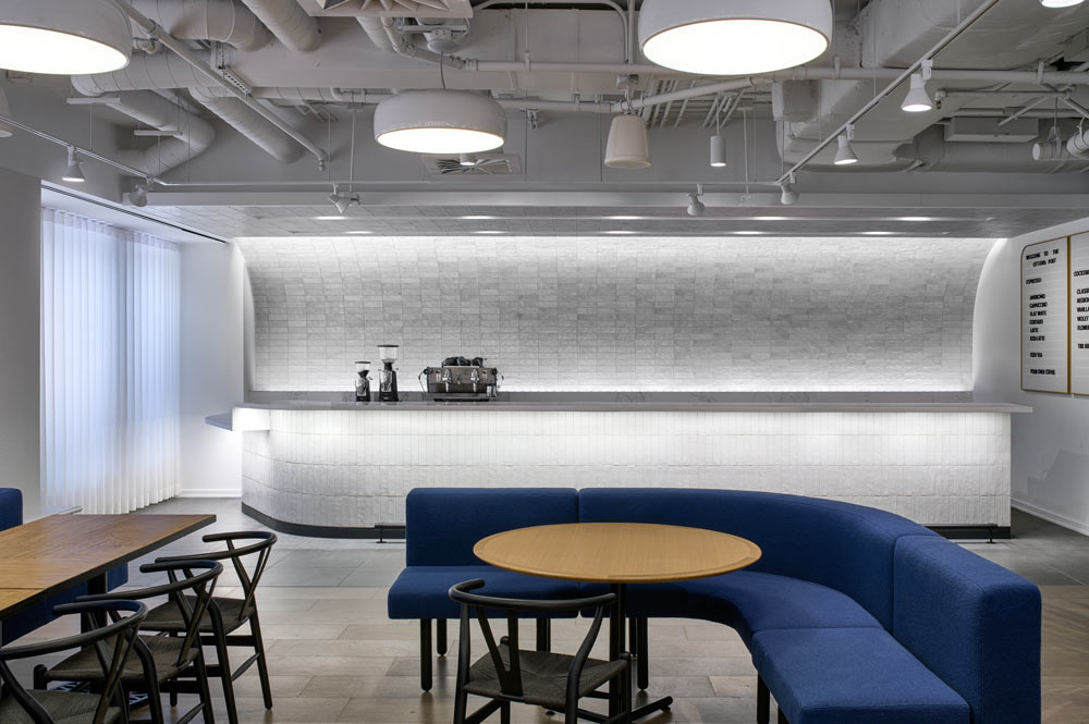 Coffee shop area, in predominantly white, with furniture as accents in blue and black. A long bar dressed in small horizontal white tile, and just coffee machines displayed on the countertop