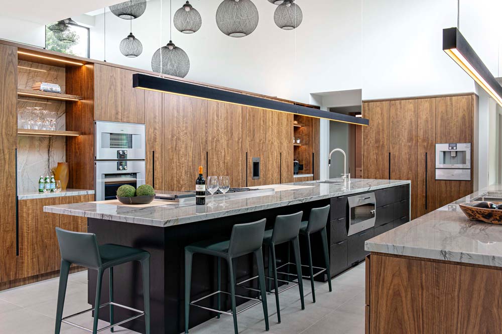 Kitchen on the main floor is mainly in wood finish cabinetry and a large island in the middle of the space. The island has a black base and modern black bar stools on one side, while clean linear light fixture hangs above it.