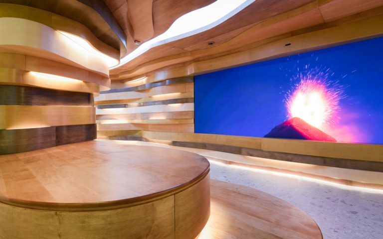 A view inside the organic occupiable space of undulating wood striations akin to natural cave formations seen in nature, with a large LED screen that displays nature imagery