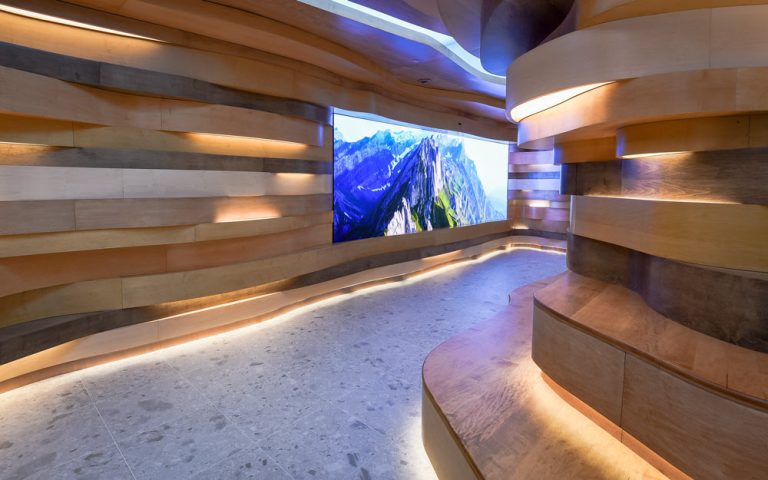 A view inside the organic occupiable space of undulating wood striations akin to natural cave formations seen in nature, with a large LED screen that displays nature imagery