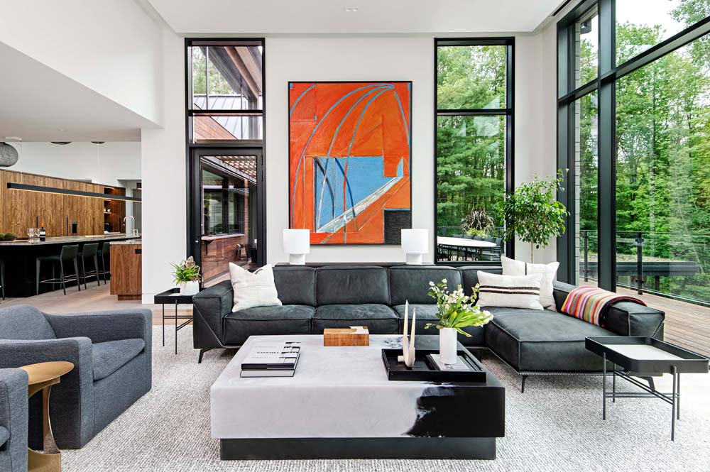 Double height living room is surrounded by floor to ceiling modern windows and forest views, while modern dark seating furniture and large square coffee table adorn the space. The focal point is a colourful piece of art hanging on the wall above the sofa.