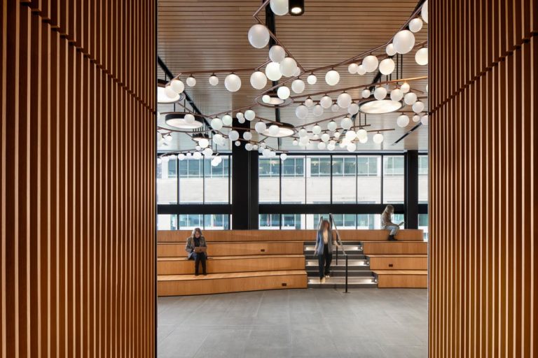 A view through a hallway with slatted walls on both side, toward wooden tiered seating is inspired by the topography of Mount Royal and above is the bubble-clustered chandeliers