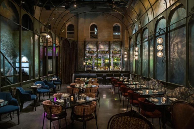 This main are of the restaurant shows custom whimsical murals framed by a custom Victorian greenhouse, a tent-draped mezzanine and a 2-story mirror installation. On the left is the stair case to the mezzanine and at the back of the room is the bar with tall shelves displaying bottles