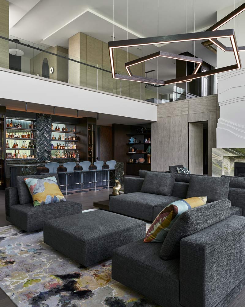 Double height living room with ample seating like couches, sofas and chairs and geometric light fixture above. A large bar is in the back of the room