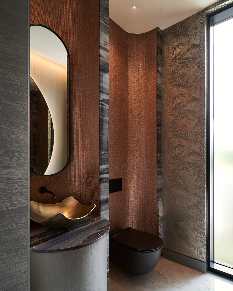Warm toned bathroom with luxurious wall coverings, black modern toilet and a golden unusually shaped sink and oval mirrior