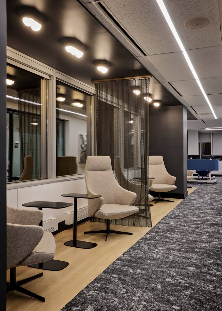 A waiting/lounge area in a long hallway with neutral coloured lounge chairs, and black detail running along the ceiling above, leading to the cubicle area