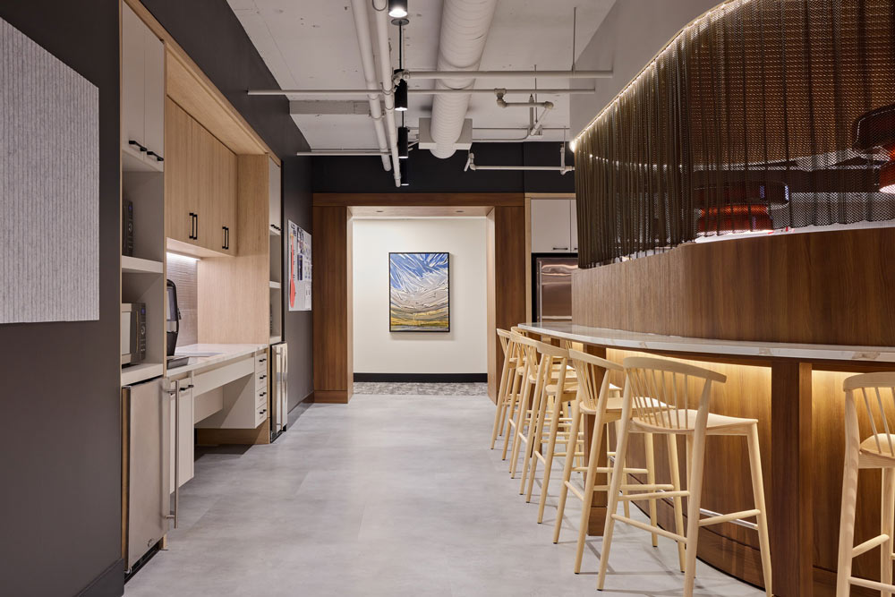 Kitchen and bar area featuring high loft-like ceiling, with a sleek wooden bar with stools on the right side and kitchen cabinetry on the left.