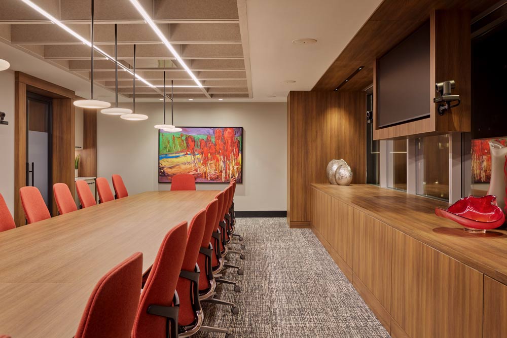 A board room with a long table in the centre and red chairs, and a built in wall cabinet along the wall housing a screen. At the end of the table is a colourful abstract painting