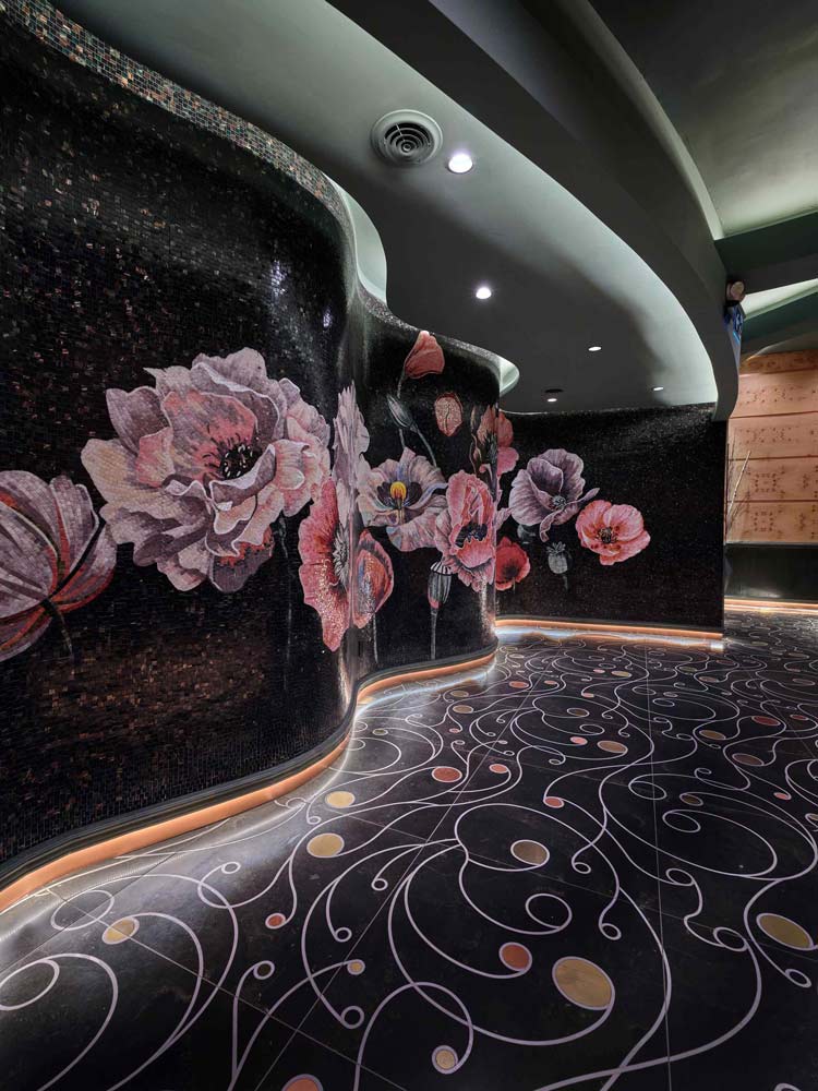 A 100-foot-long curvilineal wall of custom encrusted porcelain mosaic tiles, expressing a bold floral garden, lead the eye to the reception desk. Custom designed and fabricated black ceramic tile floor, with free-flowing curved inlays of brass and copper