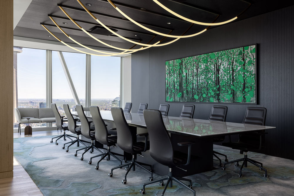 A board room with a long table and multiple rounded linear lighting rods hang above. The wall in the back is black panelled wall with a large picture of the woods.