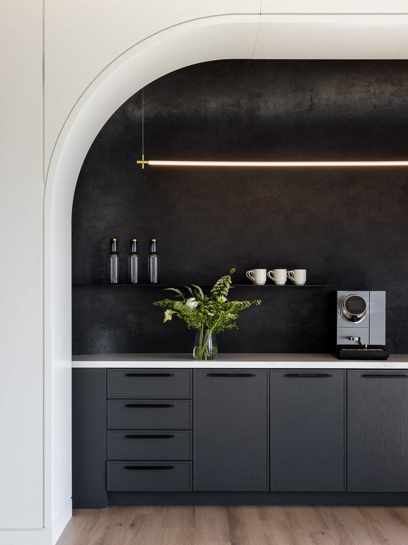 The black minimalistic kitchen area has a thin linear light fixture above, and is enveloped in a clean white arch. A vase with green flowers is on the kitchen counter