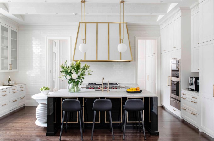 Large spacious kitchen in white finishes except the black base of the island. A round marble counter for working with dough and pastries. A custom hood manufactured from bronze and back painted glass sits over a large stove