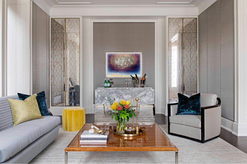 Living room space in neutral tones with a few colourful accents, contemporary sofa and a lounge chair across it with a square wooden coffe table in the middle. In the wall behind are symmetrically positioned vertical glass Eglomise surfaces - glass with mirror effect
