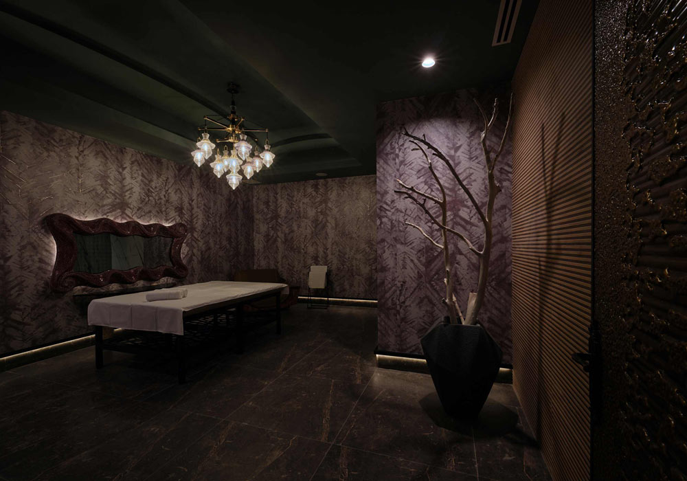 A dark and moody massage room in a grandiose luxury aeshtetic with patterned wall coverings, and wood detailing, dark coloured ceilings. A luxurious multi light chandelier hangs above the massage table