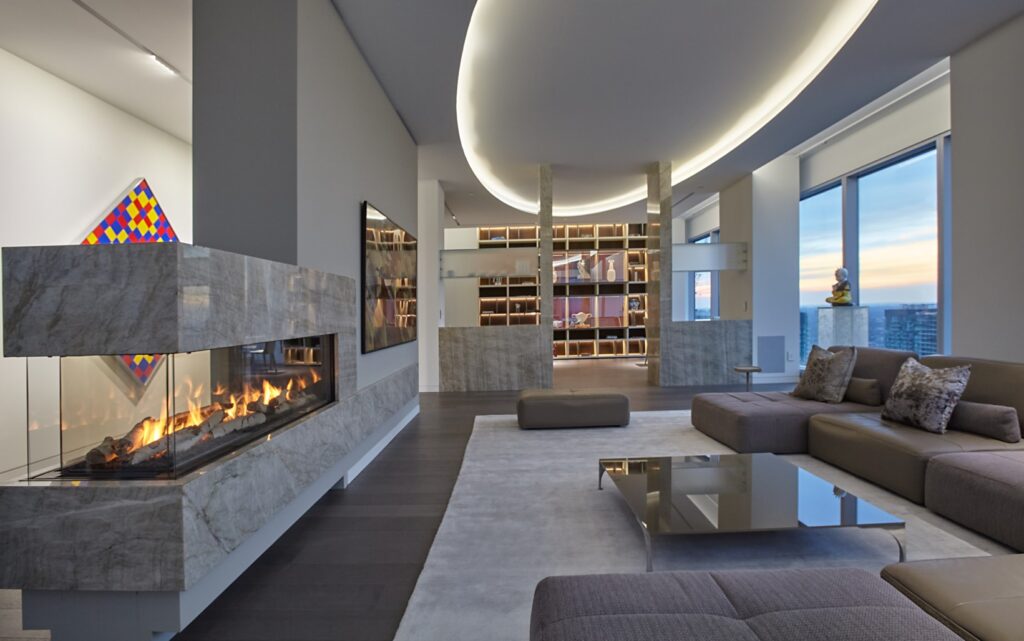 This is the second living room with a double sided fireplace in a wall separating the foyer from the room. Modern large sectional furniture is in the centre and in the very back a large builtin shelf is visible