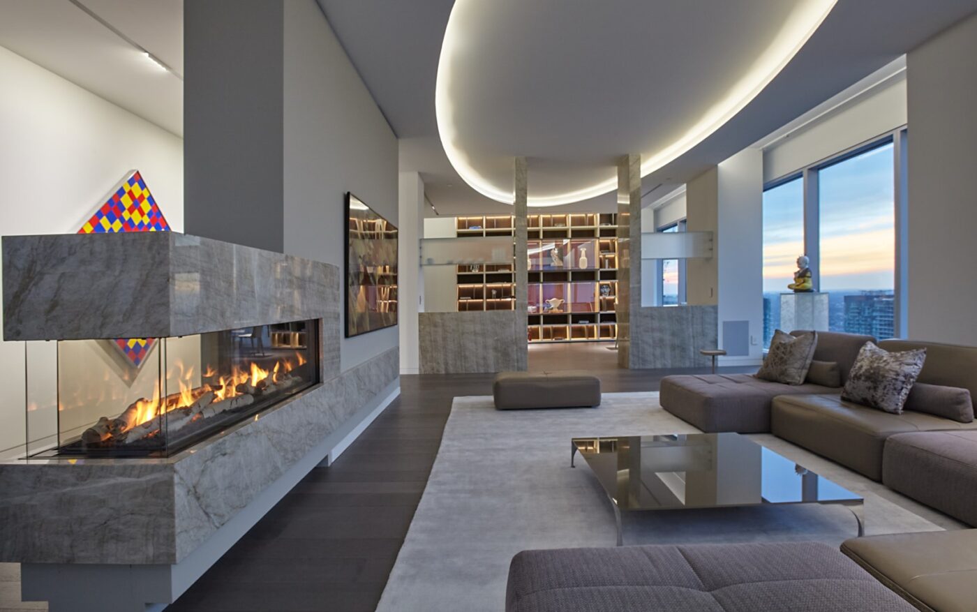 This is the second living room with a double sided fireplace in a wall separating the foyer from the room. Modern large sectional furniture is in the centre and in the very back a large builtin shelf is visible