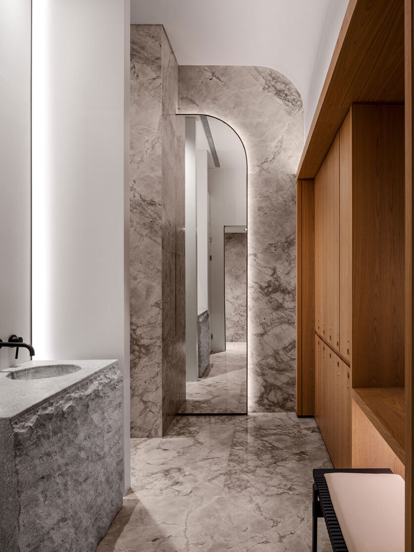 Gym and Spa washroom in predominantly marble finish with wooden lockers on the one side and stone based sink on the other. In the middle is a curved edge backlit mirror