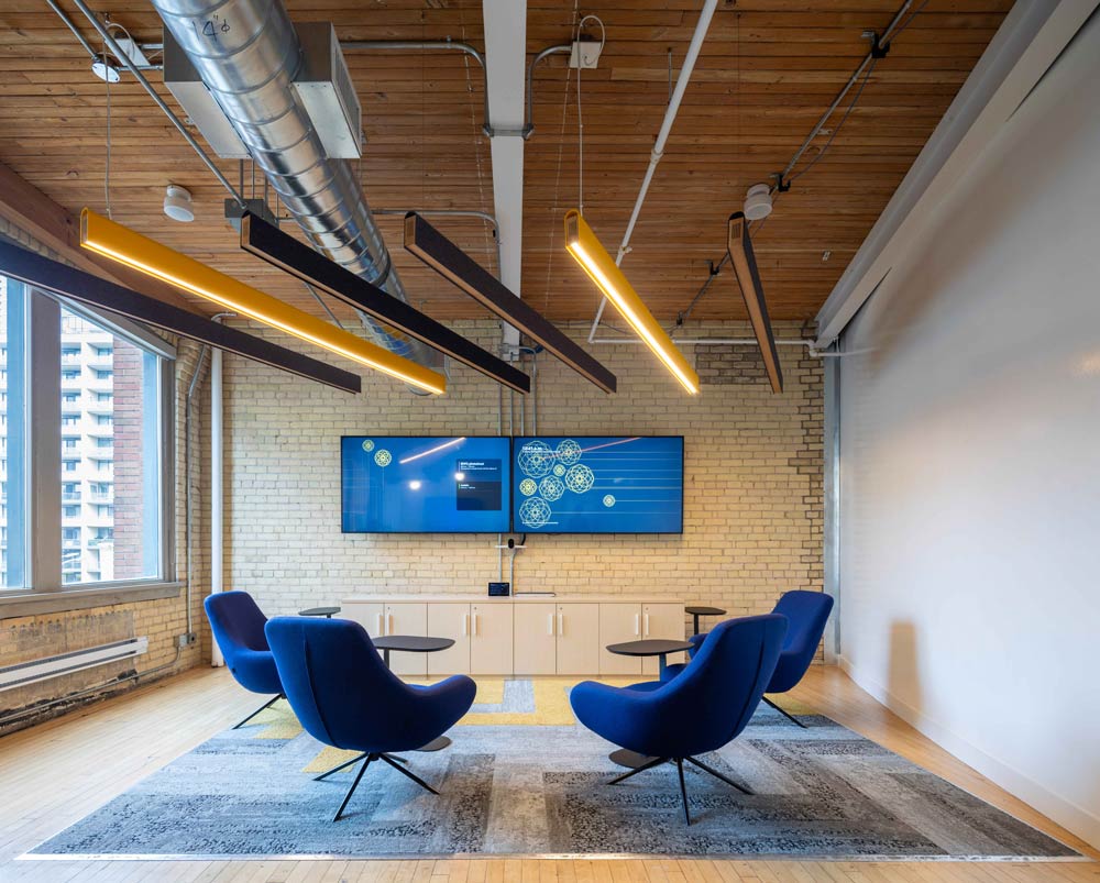 Four lounge chairs with small side tables all pointed toward a large screen on a brick wall. Above are linear yellow and black lighting hanging light fixtures