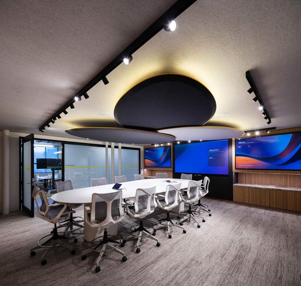 Cockpit room, a boardroom with a large white oval table and chairs, and three black acoustic oval panels above on the ceiling. On the wall beside the table are three large touch enabled TV screens.