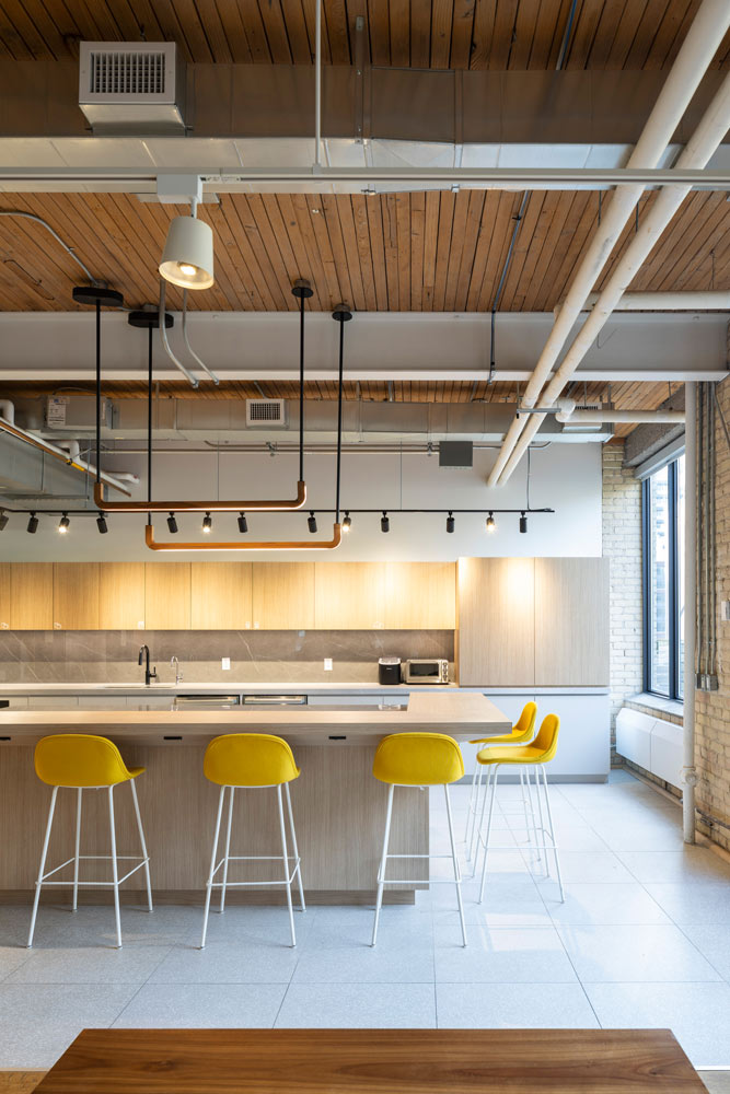 Large communal kitchen area, in neutral tones, yellow bar stools at the island, and exposed beams and hvac above that add an industrial feel.