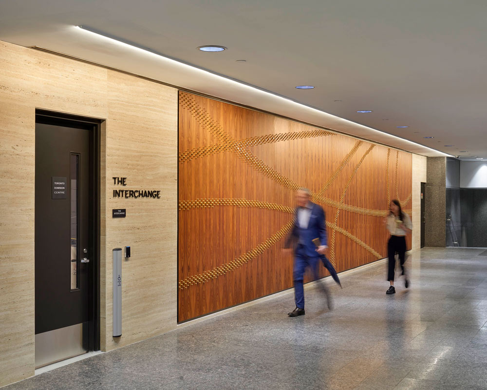 People walking by the Interchange Mural that's on the wall made up of brass pegs across a wood finished panel
