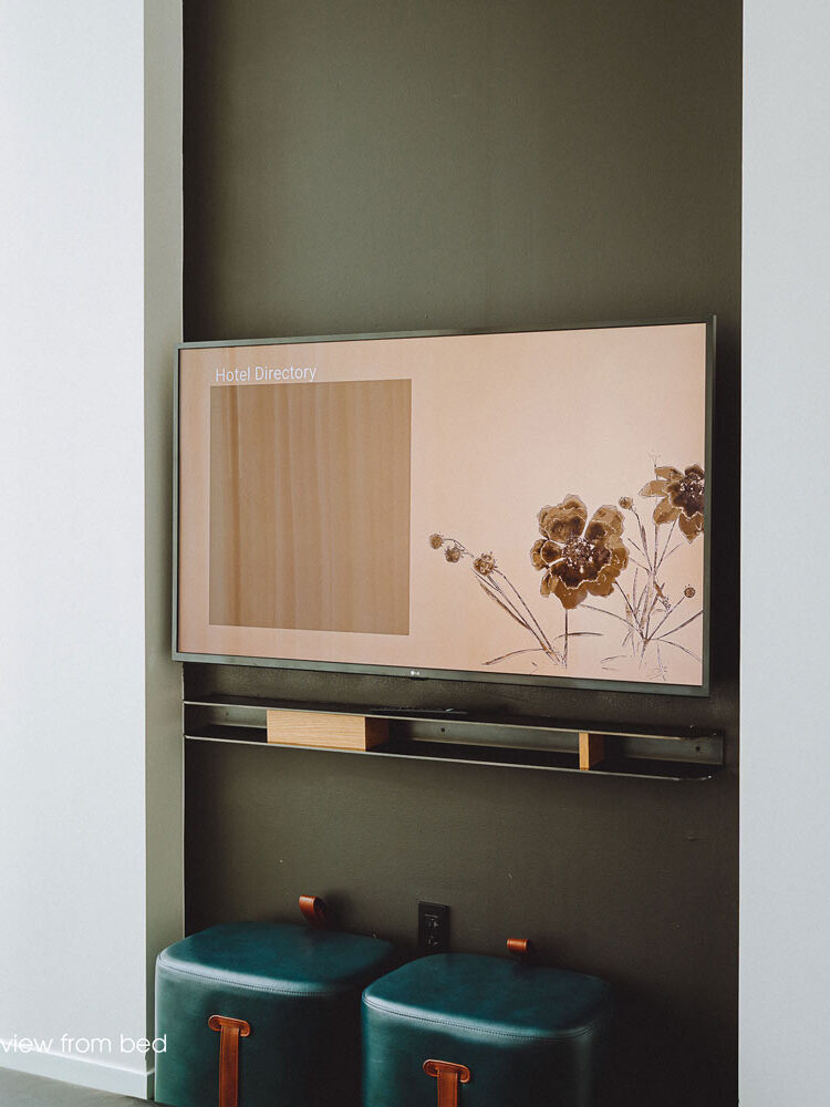 View from the bed on to the opposite wall with a flat screen tv hanging on a dark green accent wall, with two small green ottomans underneath