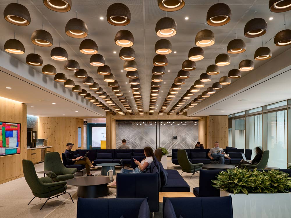 large common lounge/work area with lounge seating in dark blue and green, lit by numerous round pendant lights arranged in linear pattern leading the eye toward the back of the interior