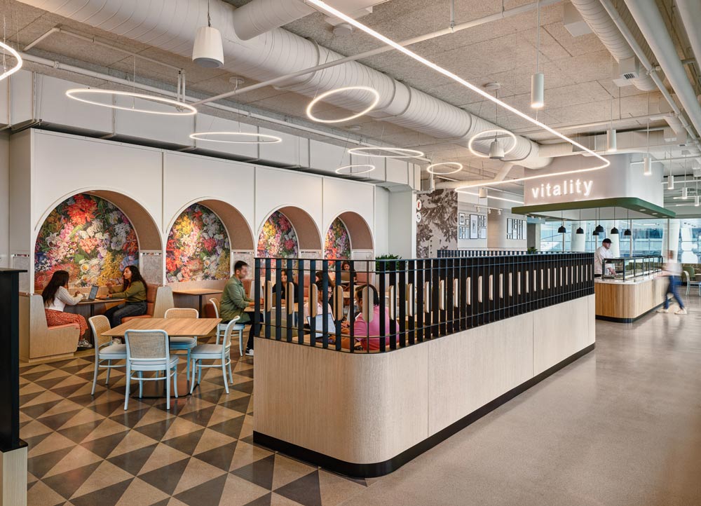 Cafeteria area enclosed between a mid-height privacy wall on one side and on the other are 4 arched booths with colourful floral wall paper on the wall. The floor features a geometric triangular black and white pattern
