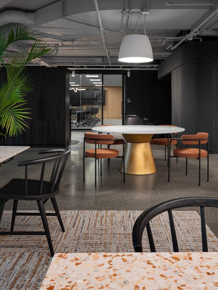 Round table with golden finish geometric base and a large white light pendant above, in a large open space in mostly black and dark colour scheme