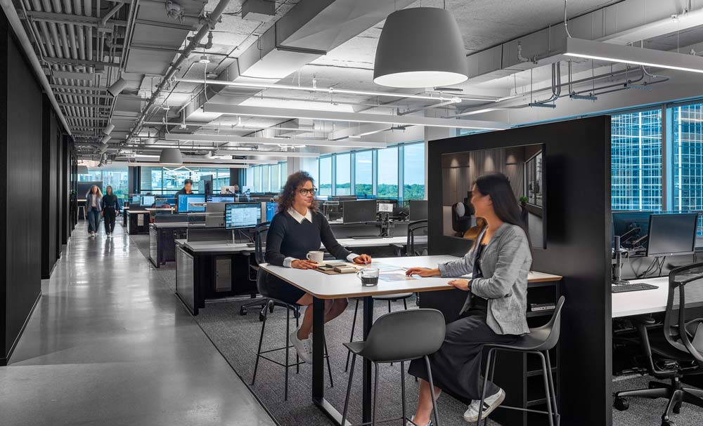 Work station area with rows of desks across the space in predominantly black and grey colour scheme, industrial unfinished ceilings. In the forefron are two women sitting at a tall table, having coffee and talking