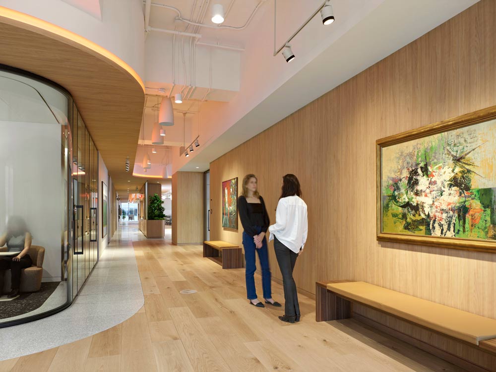 Two women standing in A wide and spacious hallway, in light wooden finishes on the floor and wall where a large art work hangs