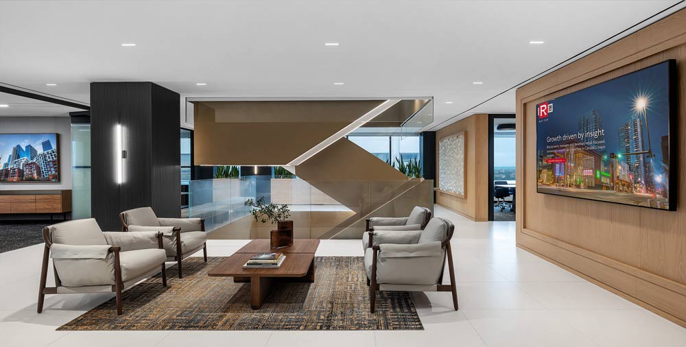 Lounge area with four lounge chairs and a wooden table and behind it is a sleek modern staircase with glass railings. A large TV screen hangs on the wood covered wall to the right of the lounge area