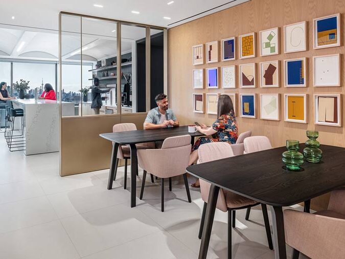 Lunch room area with tables meant for socializing and collaborating with colourful gallery wall above. The area is separated by a glass wall from the kitchen area