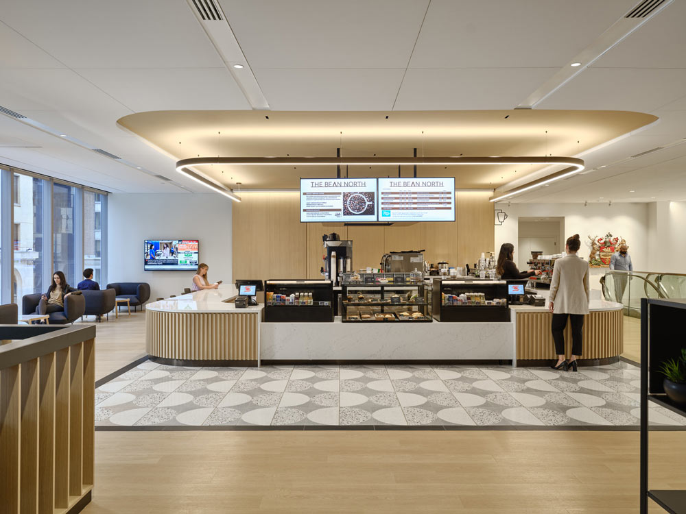 Coffee shop positioned centrally in the space, in neutral bright finishes, linear lighting above and lots of natural light coming from the windows on the left of the coffee bar