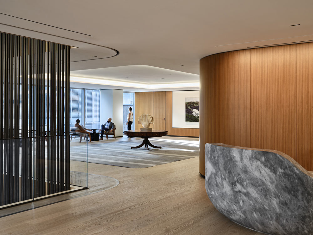 View of a central foyer with a round table and sculpture in the centre, through a narrow passage between a rounded wood covered wall and a black slatted detailing on the opposite side