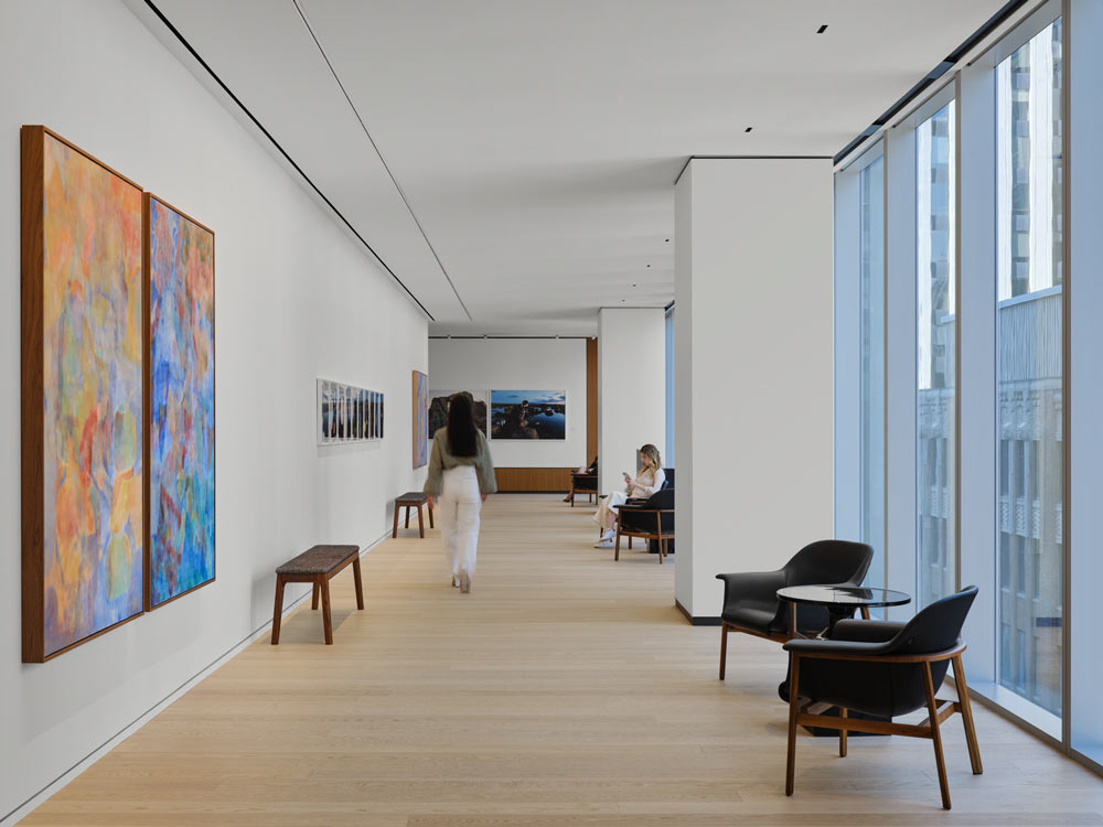 A woman walking through a wide hallway with beautiful colourful large art works on the wall on the left, and floor to ceiling windows on the right and black lounge chairs beside the windows