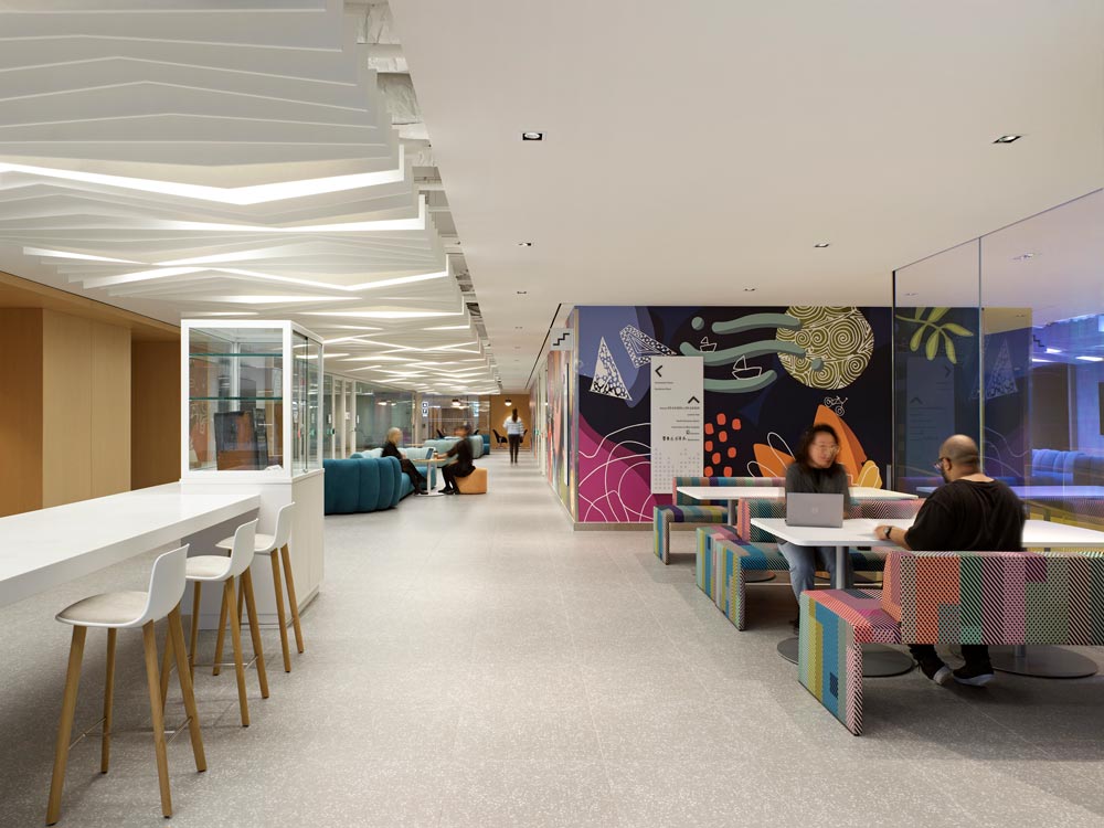 This is a workspace area with a colourful mural on the wall, colourful bench seating and interesting accordion-like lighting running along the ceiling above the hallway leading to the other side