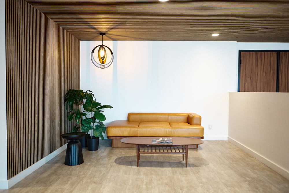 A lounge/waiting area in the lobby with a yellow leather sofa and mid century stule coffe table and side table. The wood slat detailing is on the wall and running up the whole ceiling, where a round pendant hangs above the lounge area