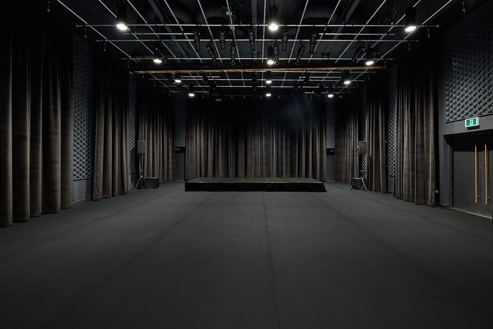 A completely black room with a stage at the far end, black floor to ceiling curtains, black floor and ceiling, and black wall coverings