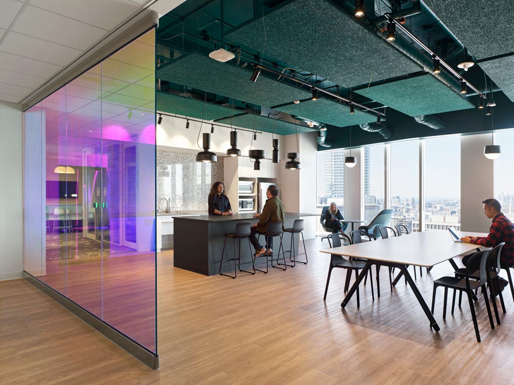 Work space with a small kitchen, dark coloured high loft style ceiling and large desk with multiple chairs. The space overlooks the city through floor to ceiling windows. A coloured glass creates a separation between the workspace and the hallway.
