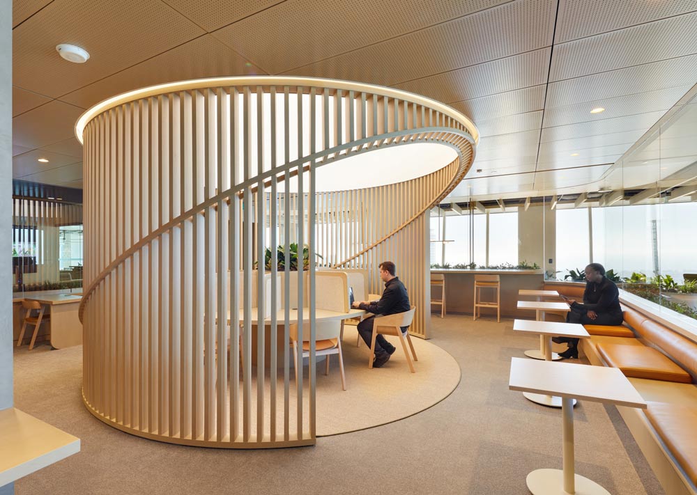 A round table divided into several small workstations is surrounded by a round privacy wall made of wood slats. All around this large round work area are benches and small tables along the glass walls looking onto the the hallways