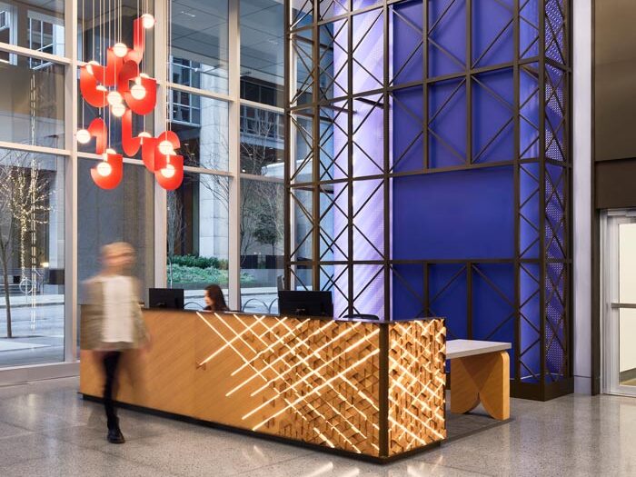 Reception desk and behind it is a bright blue/purple wall with metal bars across it in a geometric pattern formation. A modern bright red light fixture hangs, made up of multiple cascading geometric shapes